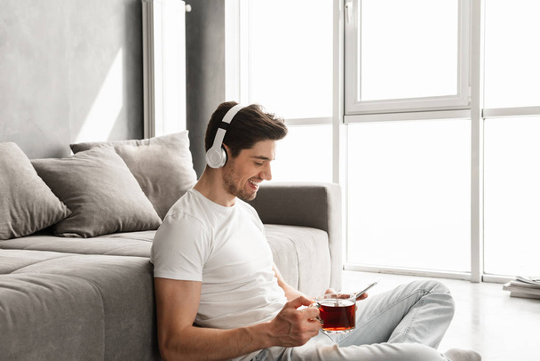 Photo in profile of young guy listening to music on smartphone wearing wireless earphones while sitting on floor with cup of tea in hand - Valokuva, kuva