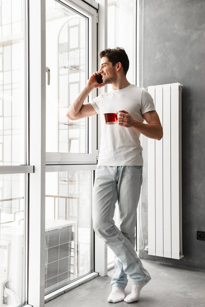 Full length photo of joyful brunette man in casual wear speaking on mobile phone and looking out the window while holding glass with tea - Fotografie, Obrázek