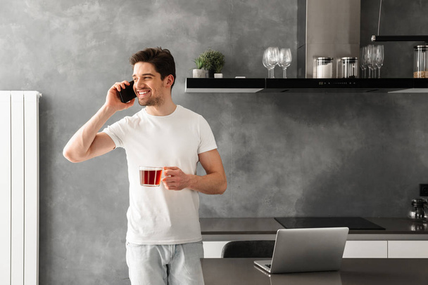 Beautiful cheerful man speaking on mobile phone and looking aside while holding glass with tea - Foto, Bild