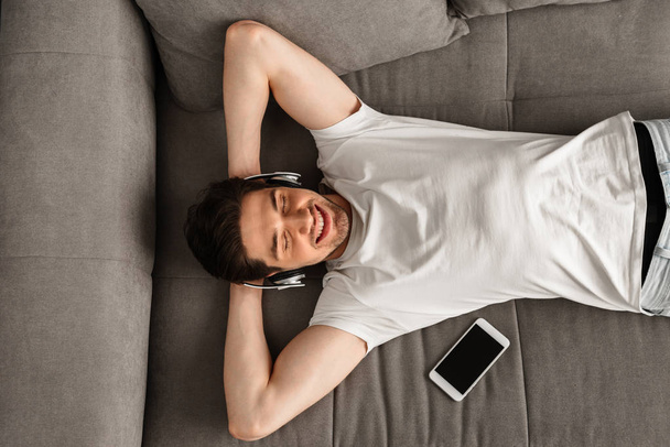 Photo from above of modern man 30s in white t-shirt lying on sofa in flat while using smartphone and earphones - Фото, изображение