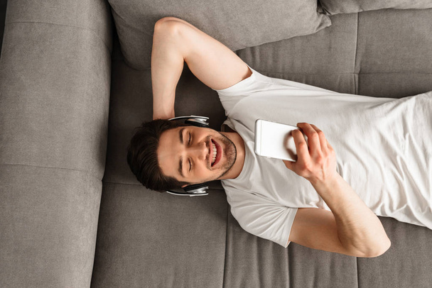 Photo from above of handsome masculine man 30s in white t-shirt and earphones lying on sofa in flat and looking on mobile phone - Valokuva, kuva