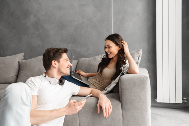 Caucasian loving couple man and woman in casual clothing resting in living room in modern flat while using laptop computer and smartphone - Valokuva, kuva