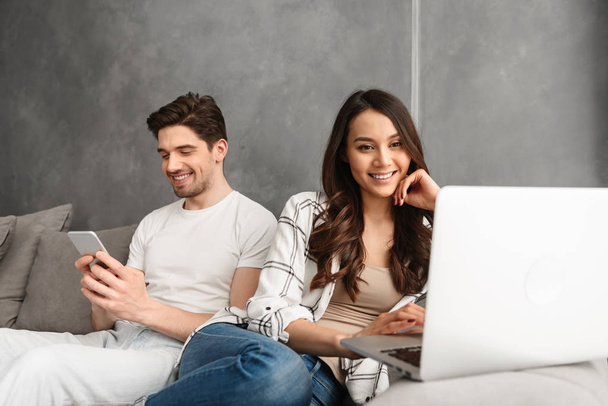 Joyful man and woman sitting together on couch in gray interior while using laptop and smartphone - Photo, image