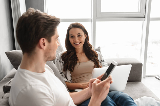 Caucasian couple man and woman in casual clothing resting in living room at home while using laptop computer and smartphone - Foto, Imagem
