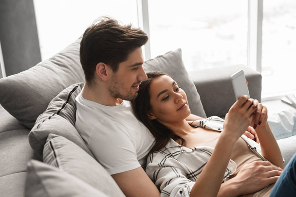 Portrait of gorgeous people man and woman lying on sofa at home and using mobile phone - Photo, Image