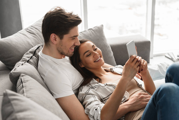 Happy family portrait of loving man and woman lying on sofa at home and using mobile phone - Fotografie, Obrázek
