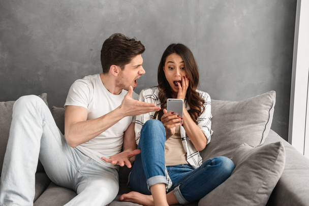 Confused woman holding mobile phone in hand with puzzlement while sitting on sofa at home and man shouting on her isolated over white background - Photo, Image