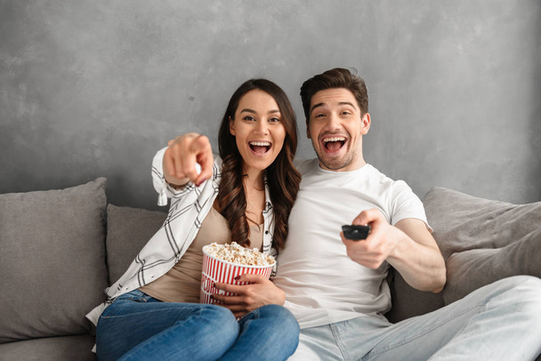 Photo of smiling couple sitting on sofa at home and pointing at you with remote control and pop corn in hands - Foto, imagen