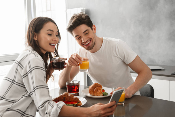 Portait of loving couple man and woman taking selfie on mobile phone while having breakfast in kitchen - Photo, image