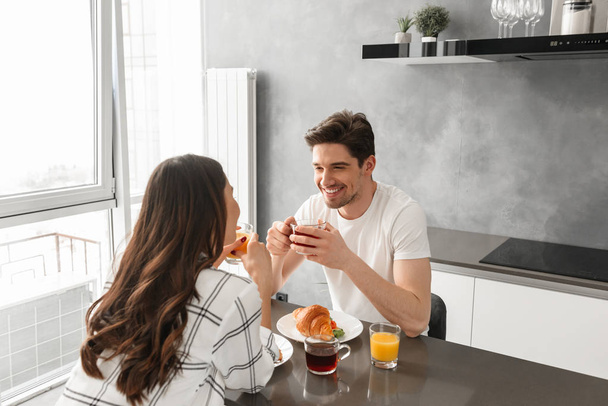 Portait of pleased couple looking at each other and smiling while sitting in kitchen with food on table - Φωτογραφία, εικόνα