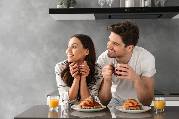 Young and beautiful couple looking aside and smiling while having breakfast and drinking tea in flat - Photo, image