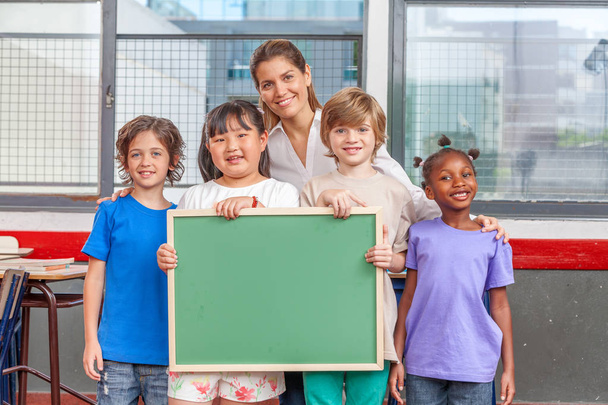 Πολυ εθνοτικών κύρια τάξη κρατώντας πράσινο πίνακα κιμωλίας, τα παιδιά και το δάσκαλο. Μετανάστευση και ένταξη έννοια. - Φωτογραφία, εικόνα