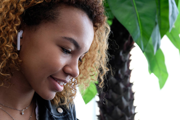 Menina afro-americana sorrindo, olhando para baixo. Retrato de uma bela jovem mulher negra moderna, em uma jaqueta de couro com airpods em seu ouvido, ouve música
. - Foto, Imagem