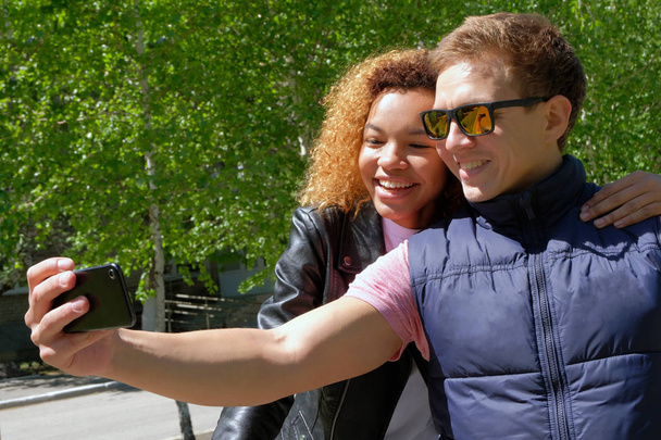 Un tipo europeo en gafas de sol con una hermosa chica de piel oscura, en chaquetas hacer selfie sobre un fondo de árboles verdes. Sonriendo a la gente en el verano al aire libre
. - Foto, imagen