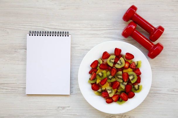 Salade de fruits frais avec haltères et cahier, vue de dessus. Pose plate. Espace de copie
. - Photo, image