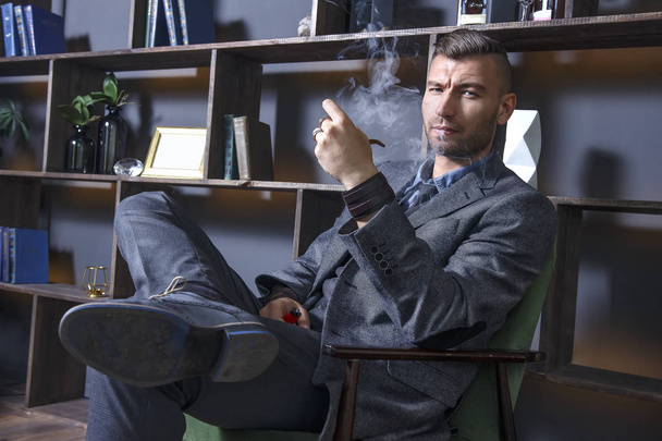 elegant man in suit sits in chair and smokes pipe. handsome stylish rich brutal man in luxurious apartment. Serious view of successful businessman. Clubs of smoke from a cigar in a dark interior. - Фото, зображення
