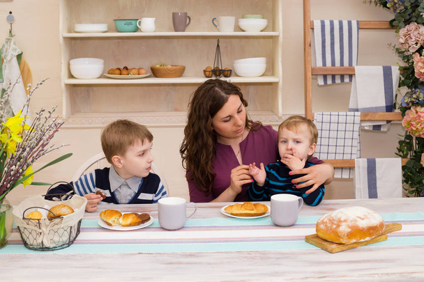 Matka i dwoje dzieci w kuchni. Szczęśliwa matka karmi jej syna. Matka i syn, śniadanie. Koncepcja szczęśliwą rodzinę i zdrowej żywności. Chłopiec jedzenia rogalika - Zdjęcie, obraz