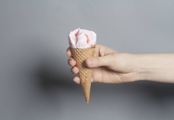 Γυναίκα χέρι κρατά ένα χωνάκι παγωτό φράουλα σε ένα γκρίζο φόντο. - Φωτογραφία, εικόνα