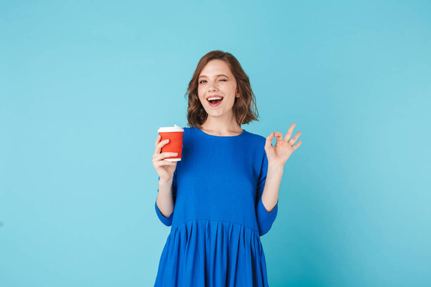 Fiatal gyönyörű hölgy dress állandó csésze kávé átmegy kék háttér. Csinos lány boldogan mutatja ok gesztus, miközben keresi a kamera - Fotó, kép