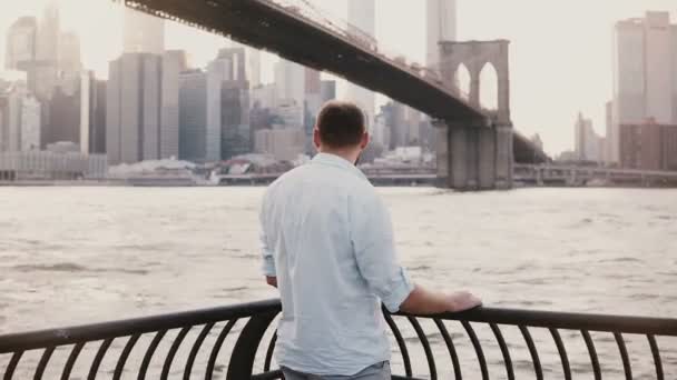 Successful happy businessman stands near Brooklyn Bridge embankment fence panorama in New York City, back view 4K. - Metraje, vídeo