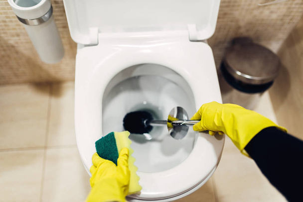 maid in rubber gloves cleaning toilet with brush, hotel restroom interior on background. Professional housekeeping service, sanitary processing - Photo, Image