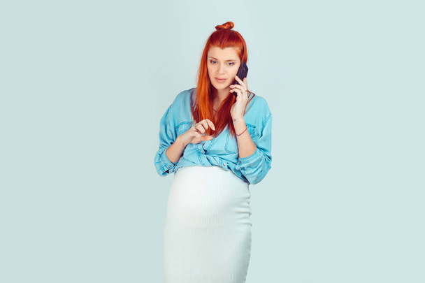 Young redhead girl expecting baby and having phone call looking away in worries isolated on light blue background. Unhappy calling her husband - Foto, imagen