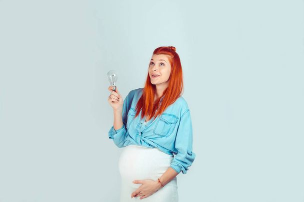 Wonderful young woman expecting baby and holding light bulb having great idea and looking up on light blue background. Good thoughts during pregnancy. Mixed race model, latin hispanic irish woman - Photo, Image
