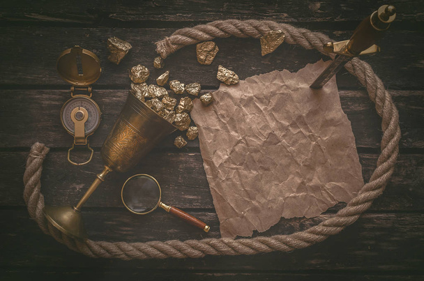 Pirate goblet full of gold and treasure map crumpled parchment with copy space, dagger, compass and ship rope on old table background. - Photo, Image