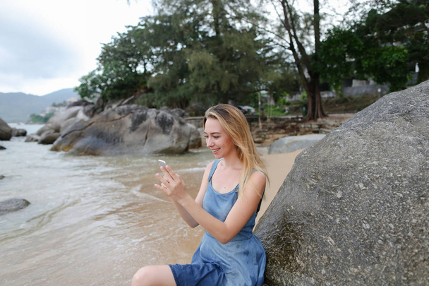 Молода жінка фотографується за допомогою смартфона і сидить на піску біля каменів і моря
. - Фото, зображення