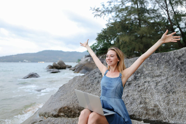Νεαρή ξανθιά γυναίκα με απλωμένα χέρια που εργάζονται με το laptop στην ακτή της θάλασσας και να κάθεται στην πέτρα. - Φωτογραφία, εικόνα