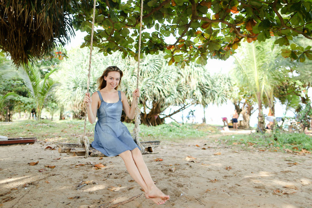 Young happy woman wearing jeans dress and riding on swing, sand in background. - Foto, Imagem