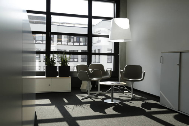 Moderno interior de oficina con muebles elegantes y lujosos. El interior es de color blanco y beige. El sol brilla a través de la gran ventana y proyecta sombras en el suelo
.   - Foto, imagen