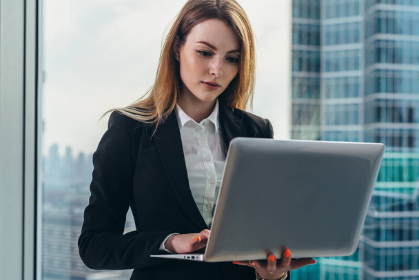 Молодая женщина-адвокат работает в своем роскошном офисе, держа ноутбук, стоящий напротив панорамного окна с видом на деловой район
 - Фото, изображение