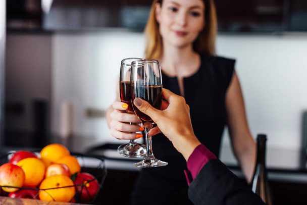 Des jeunes femmes portant une élégante robe noire tenant une bouteille de vin rouge et un verre debout au bar de la cuisine regardant son amie
 - Photo, image
