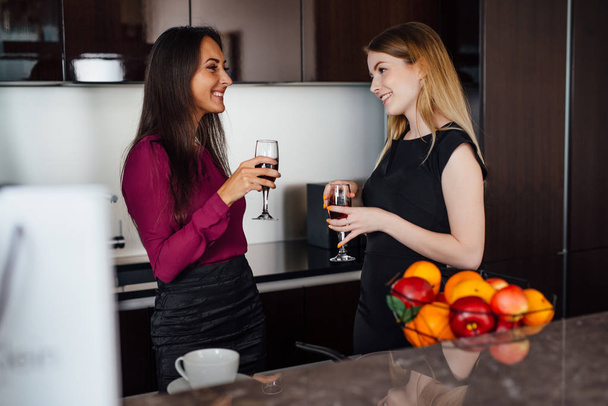 zwei Freundinnen in eleganter Kleidung entspannen sich nach dem Einkaufen, trinken Wein, lachen und plaudern in der Küche - Foto, Bild