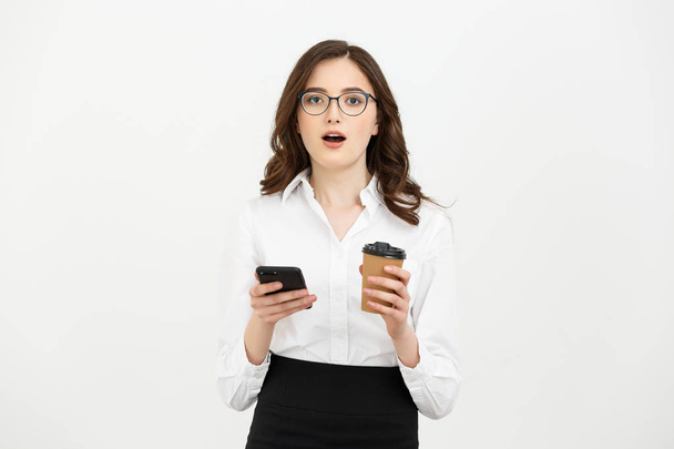 Retrato de una sorprendida mujer de negocios con anteojos que sostiene la taza de café y el teléfono móvil mientras está aislada sobre un fondo gris
. - Foto, imagen