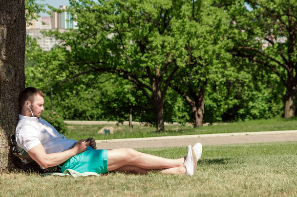Young successful businessman in a white shirt. A man is sitting on the grass, working on a smartphone in a city park on a green lawn outdoors in nature. Mobile office, business concept - Foto, Imagen