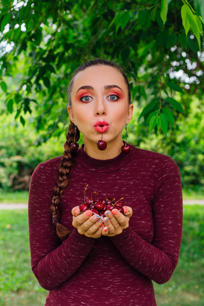 Beautiful young woman with long braid and cherry earrings holding cherry in hands and one cherry in mouth - Fotó, kép