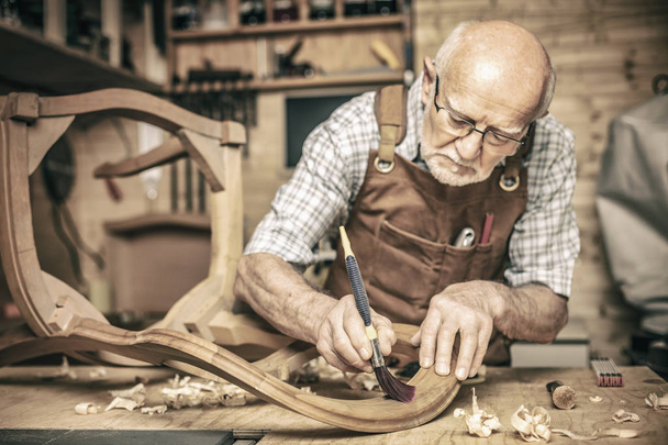elderly carpenter uses a brush on an unfinished chair - Foto, Bild