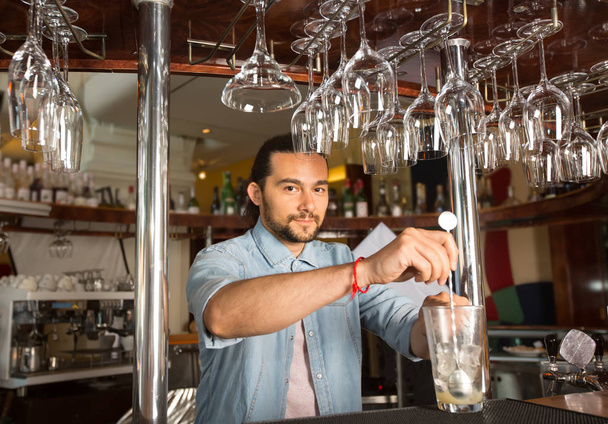 Bello giovane barista sorridente dietro il bancone del bar che tiene in acciaio cucchiaio swizzle nel vetro con ghiaccio
. - Foto, immagini