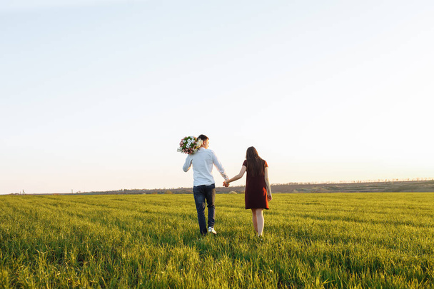 joven, feliz, pareja amorosa, al atardecer, de pie en un campo verde, contra el cielo tomados de la mano, y disfrutando los unos de los otros
, - Foto, imagen