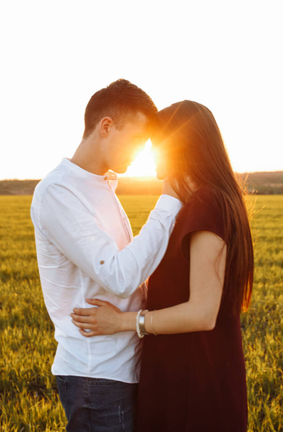 junges, glückliches, liebevolles Paar, bei Sonnenuntergang, auf einer grünen Wiese stehend, gegen den Himmel, in den Armen und einander anschauend, - Foto, Bild