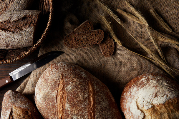 elevated view of various types of bread, knife, wicker breadbasket on sackcloth  - Fotografie, Obrázek