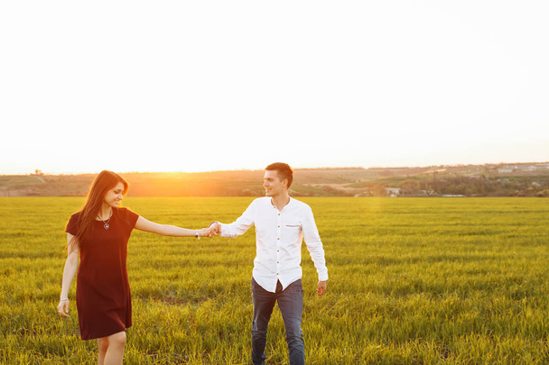 junges, glückliches, liebevolles Paar, bei Sonnenuntergang auf einer grünen Wiese stehend, Händchen haltend gegen den Himmel und einander genießend, - Foto, Bild