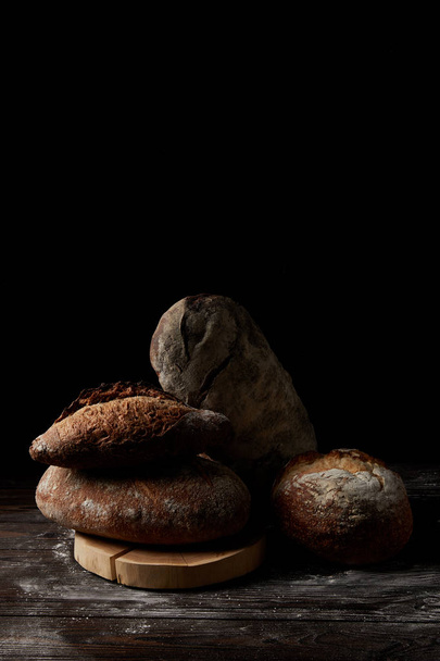 close up shot of various types of bread and cutting board on wooden table covering by flour isolated on black background - Foto, imagen
