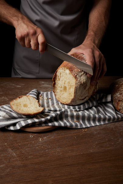 vista parcial del panadero macho cortando pan por cuchillo sobre tela de saco sobre mesa de madera
 - Foto, Imagen