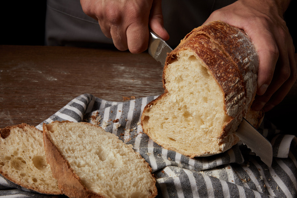 vista parcial del panadero macho cortando pan por cuchillo sobre tela de saco sobre mesa de madera
 - Foto, Imagen