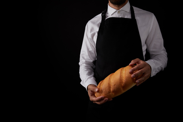 abgeschnittene Aufnahme eines männlichen Bäckers in Schürze, der Brot isoliert auf schwarzem Hintergrund hält  - Foto, Bild