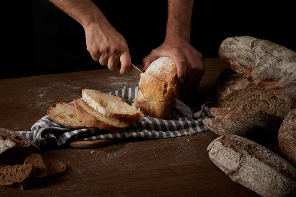 cropped shot of male baker cutting bread by knife on sackcloth on wooden table - Foto, Bild