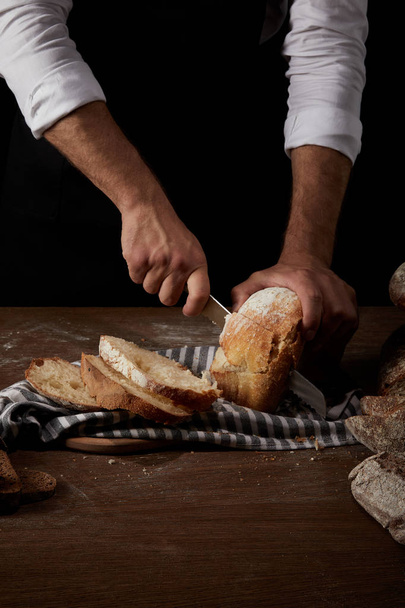 vista parcial del panadero masculino en delantal cortando pan por cuchillo sobre tela de saco sobre mesa de madera
 - Foto, Imagen